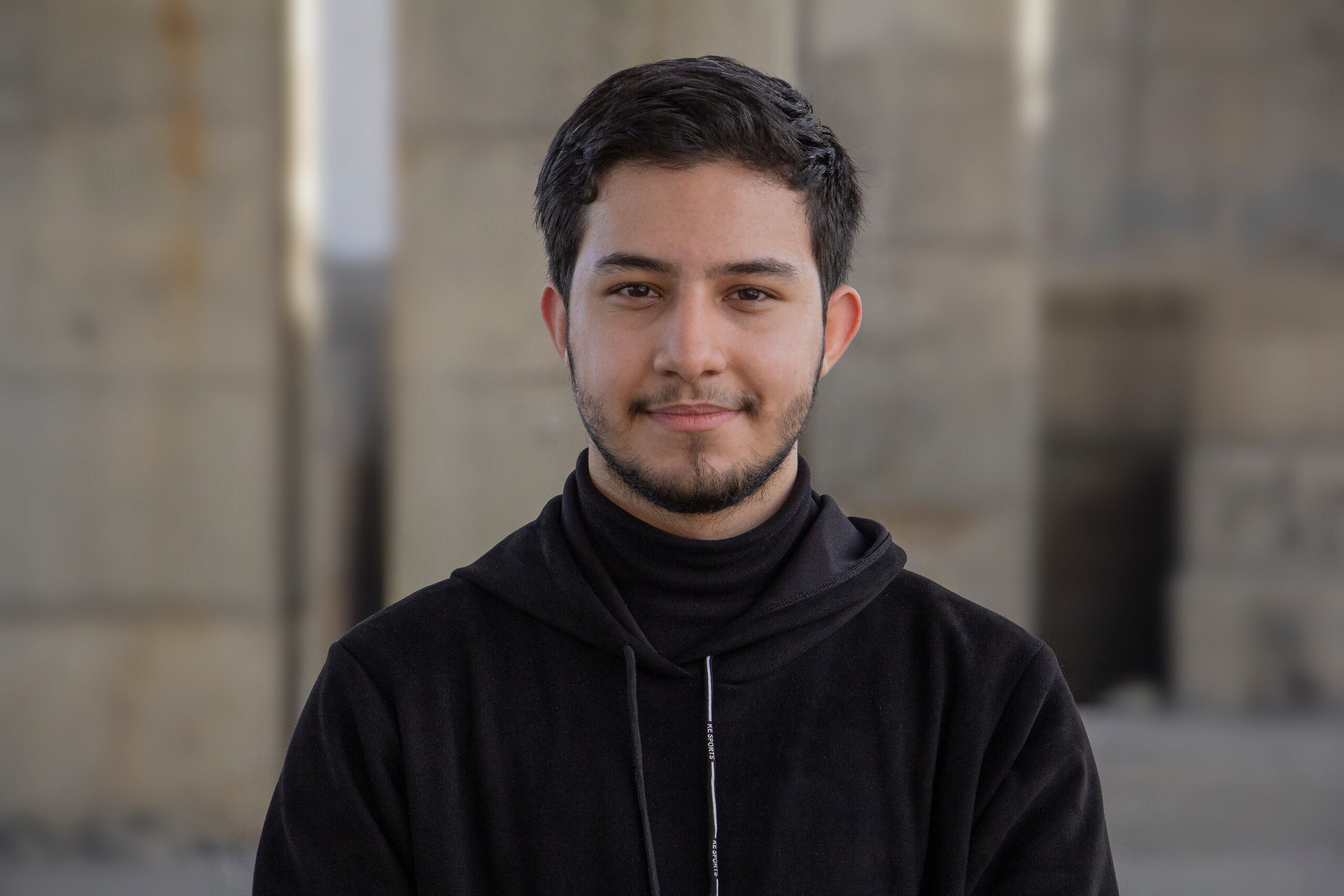 Smiling young man in a black hoodie standing outdoors, exuding confidence and warmth.